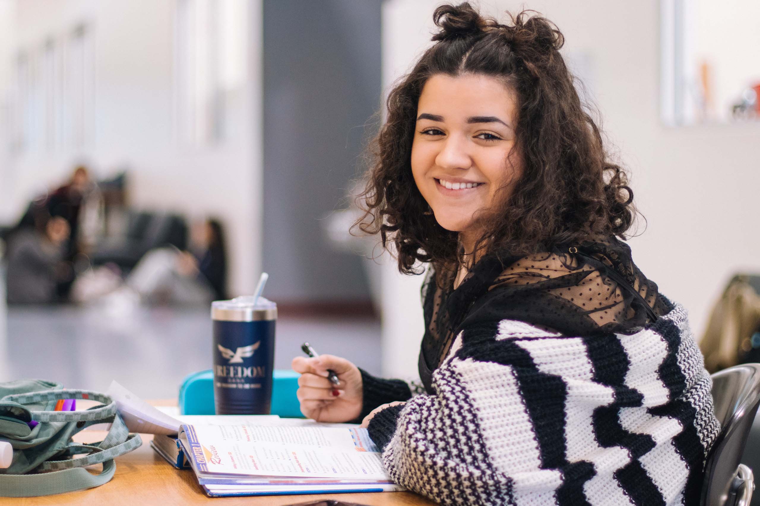 Young woman with dark curly hair and a smile looks to camera while studying