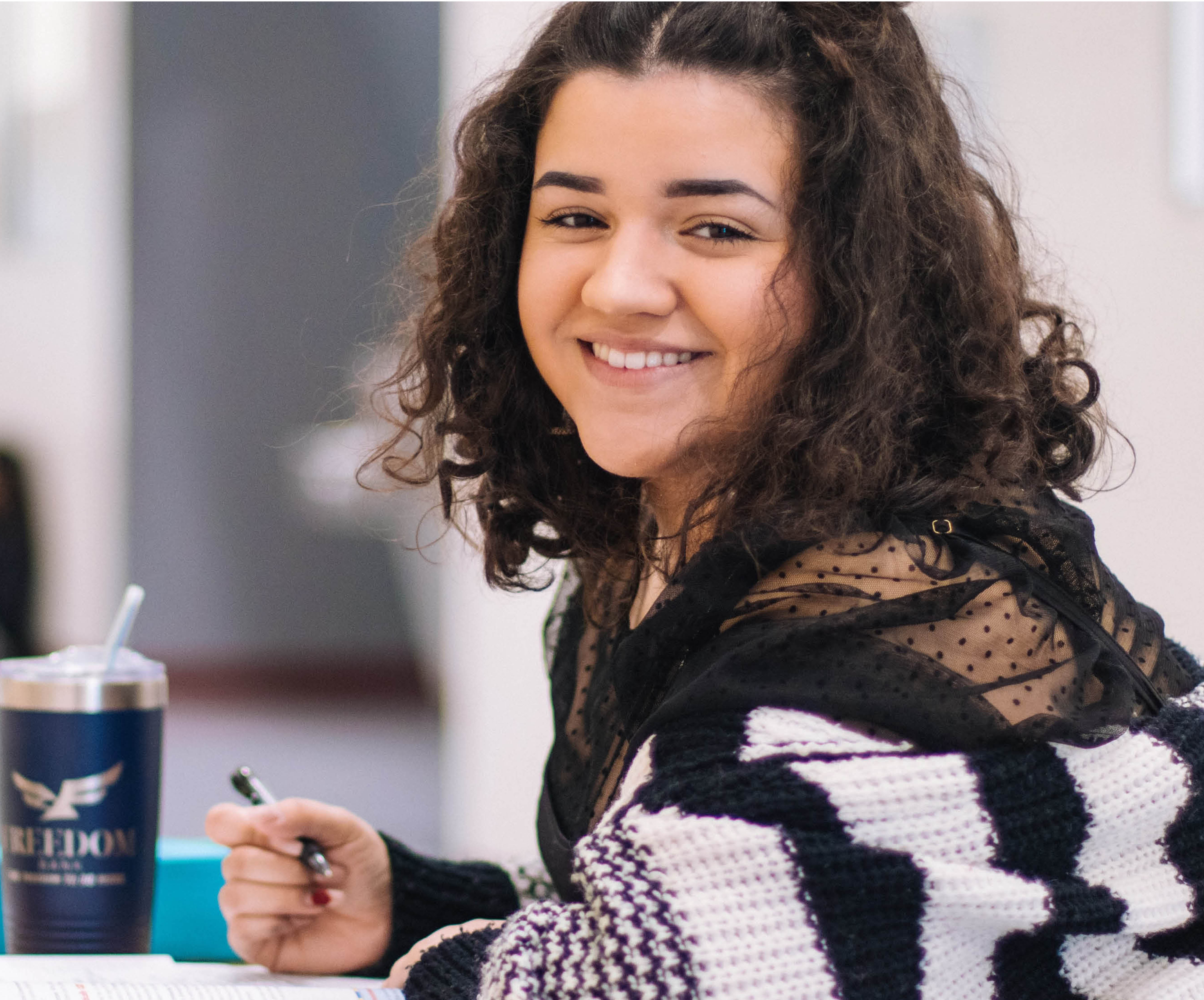 Young woman with dark curly hair and a smile looks to camera while studying