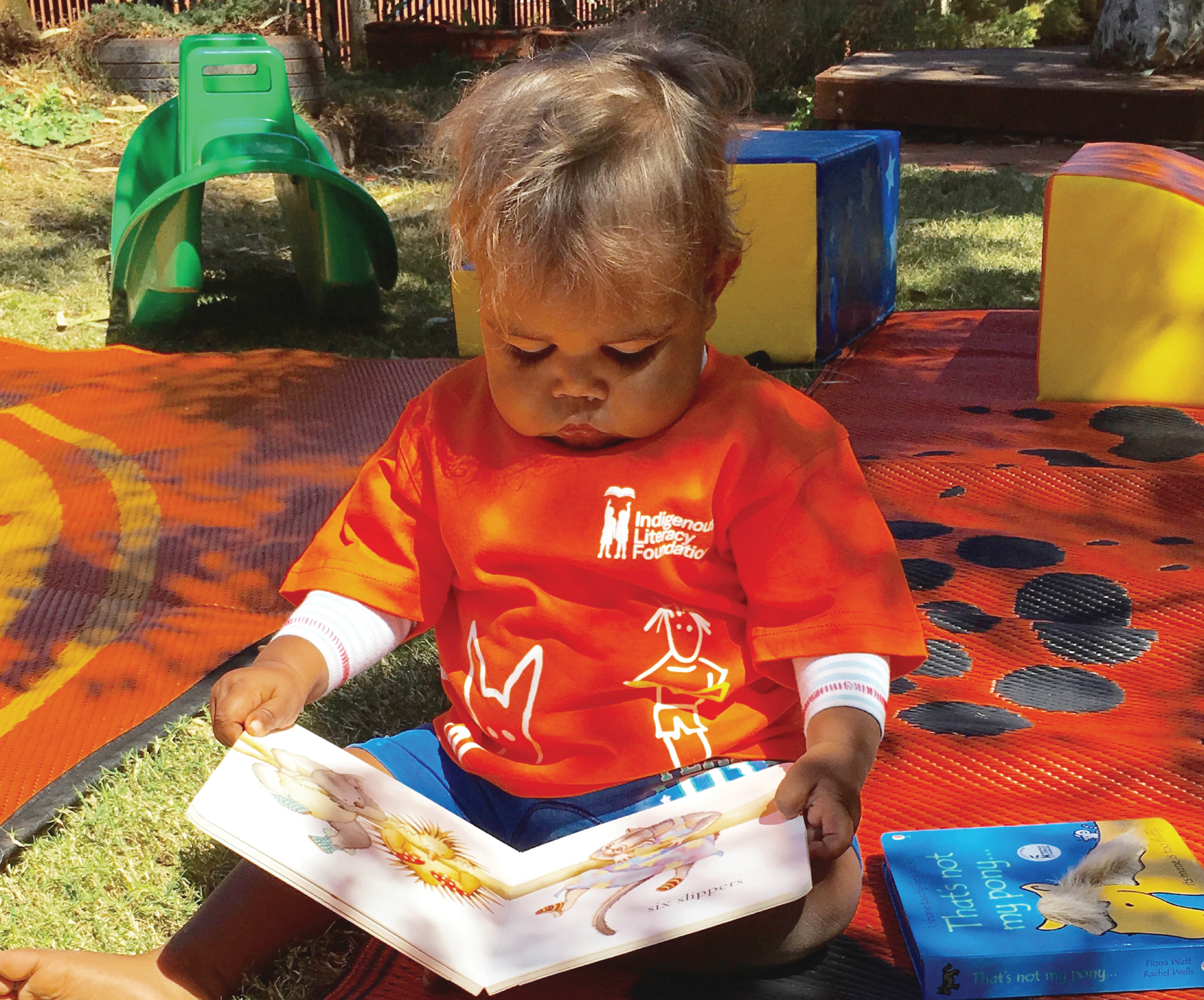 Aboriginal toddler reading possum magic board book outdoors on a blanket